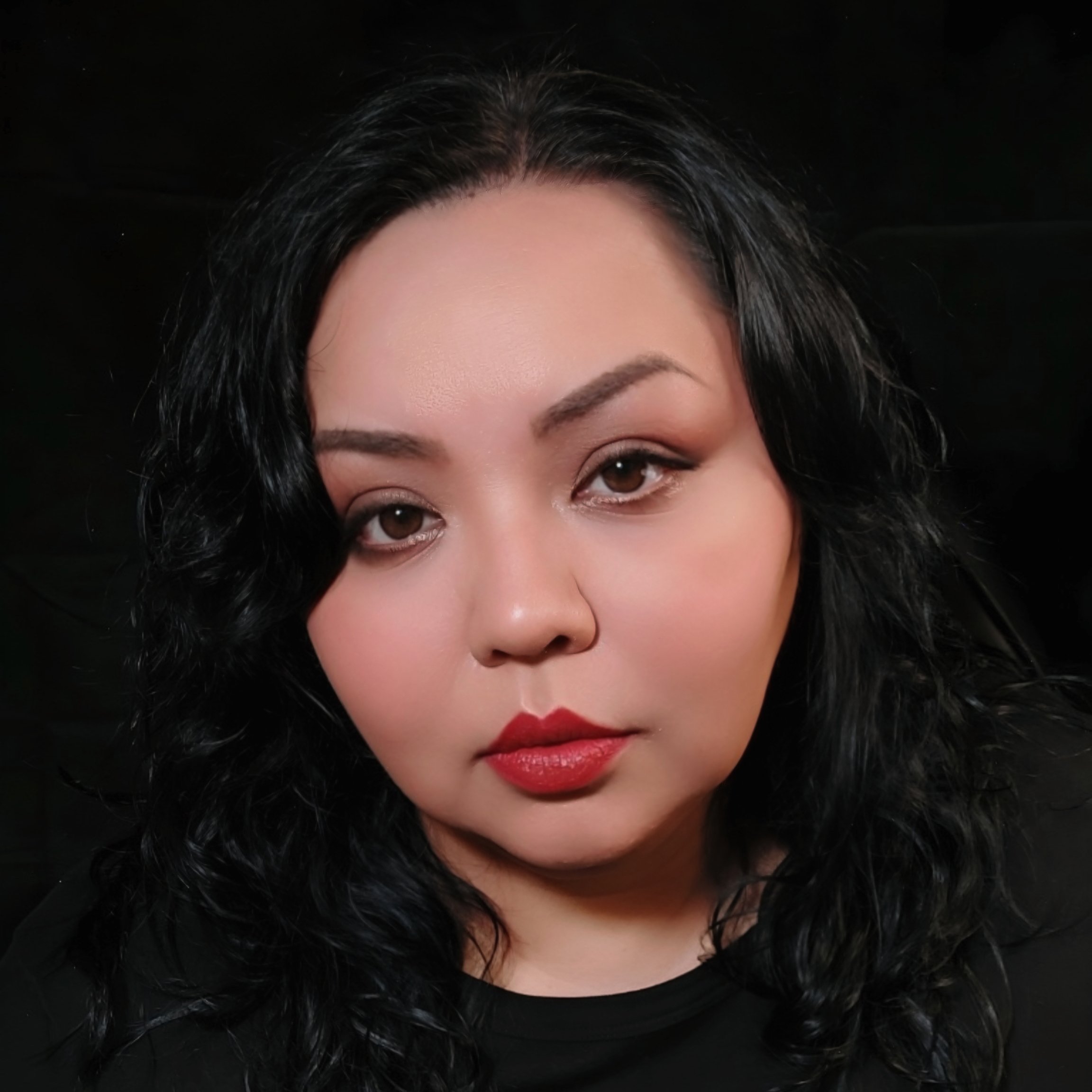 Johnnie Jae, an Otoe-Missouria and Choctaw woman, gazes directly at the camera with a slight tilt of her head. Her bright red lipstick contrasts with her black shirt, dark wavy shoulder-length hair, and black background.