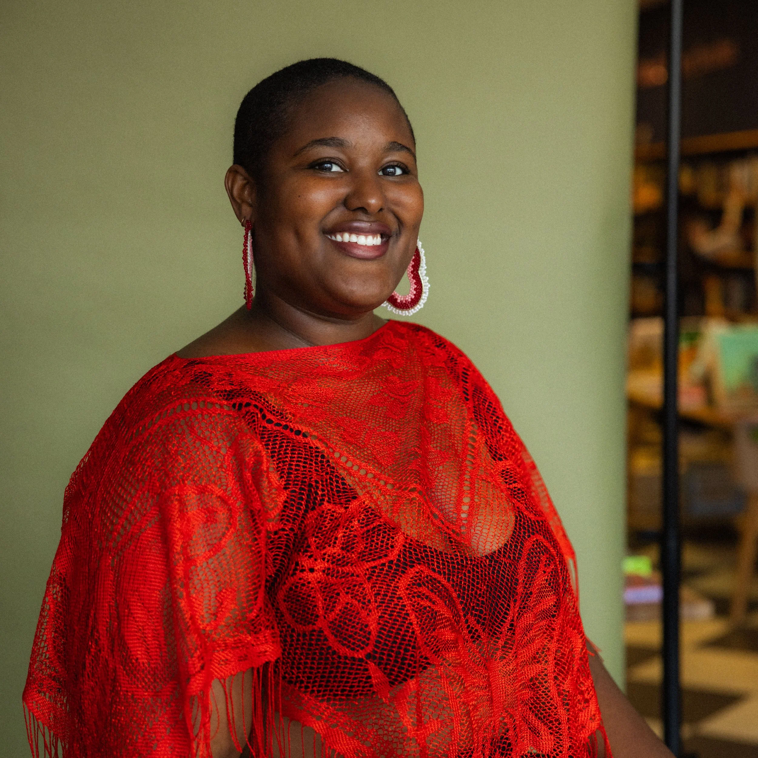 A Black person with a short haircut is seated in front of a green backdrop smiling, while wearing a red lace top and large earrings.