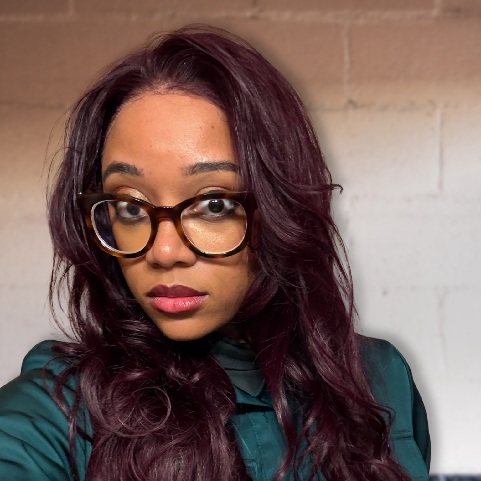 A headshot of Nadia, a Black person with deep burgundy hair, soft makeup, tortoiseshell glasses, and a silky dark green jumpsuit against a cream and teal brick wall.