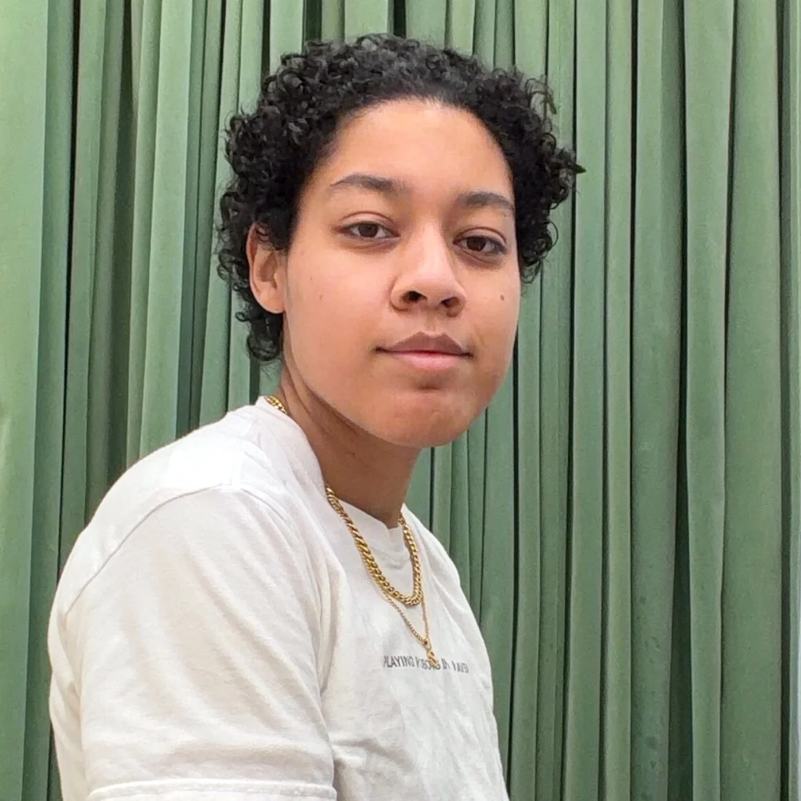 Sy’Aire, a person with short, curly dark hair, sits in front of green curtains, facing the camera with a slight smile. They are wearing a white T-shirt and a gold chain necklace.