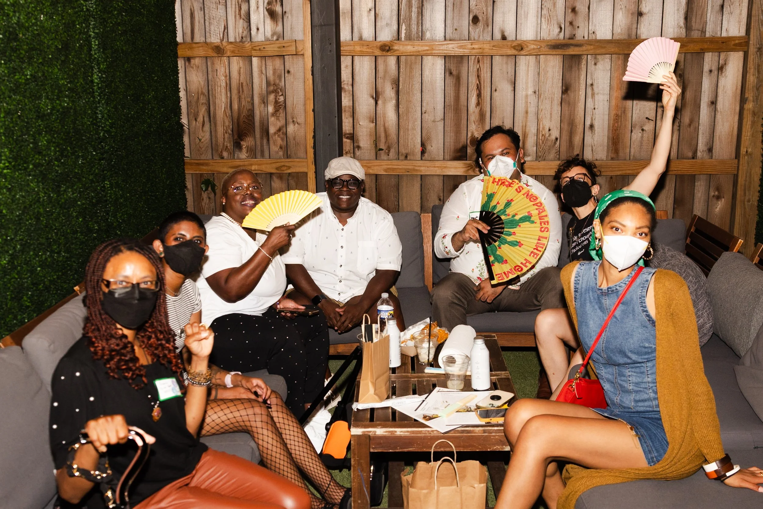 A diverse group of seven people sit together on outdoor couches at an evening event, most wearing masks and holding colorful hand fans, smiling and posing for the camera.