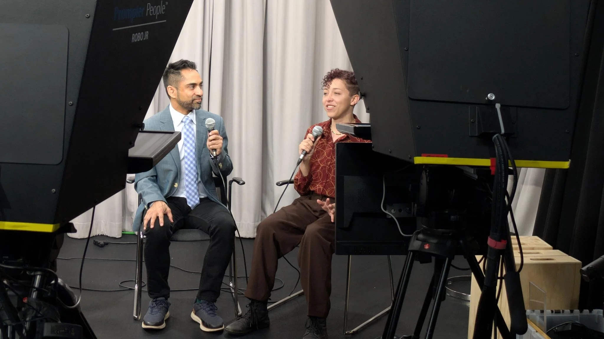 Disability Culture Lab Fellow Jaz Williams speaks into a microphone during a studio interview, seated across from another speaker. The two face each other in front of gray curtains, framed by teleprompters and production equipment.