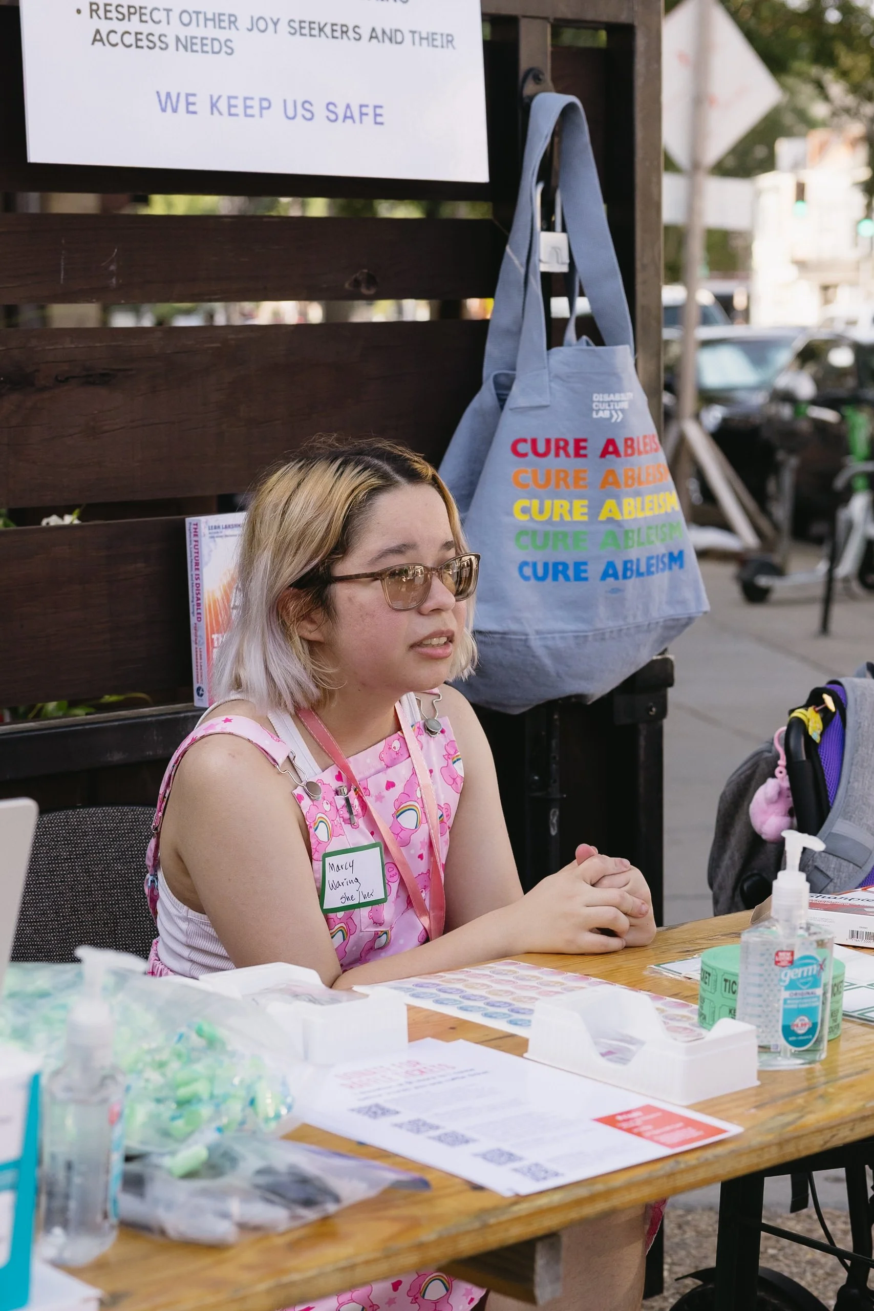 A person with shoulder-length blond hair and glasses sits at an outdoor table with flyers, sanitizer, and supplies. They wear a pink patterned outfit and name tag, with a tote bag behind them reading “Cure Ableism” in rainbow text.