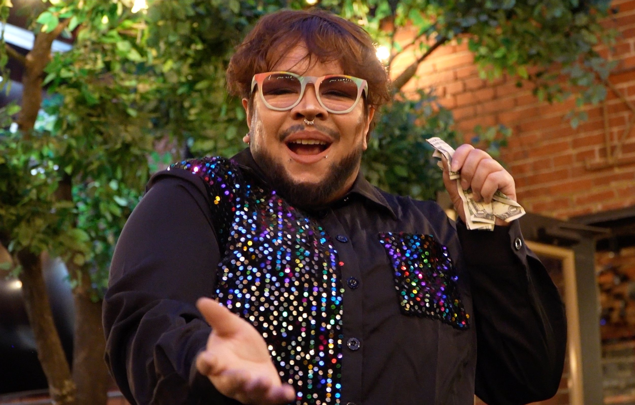 Disabled drag performer Ricky Rosé looks at the camera with an enthusiastic open mouth expression. They are wearing a black button up shirt with colorful sequins, rainbow colored glasses and a septum ring while holding dollar bills in their left hand