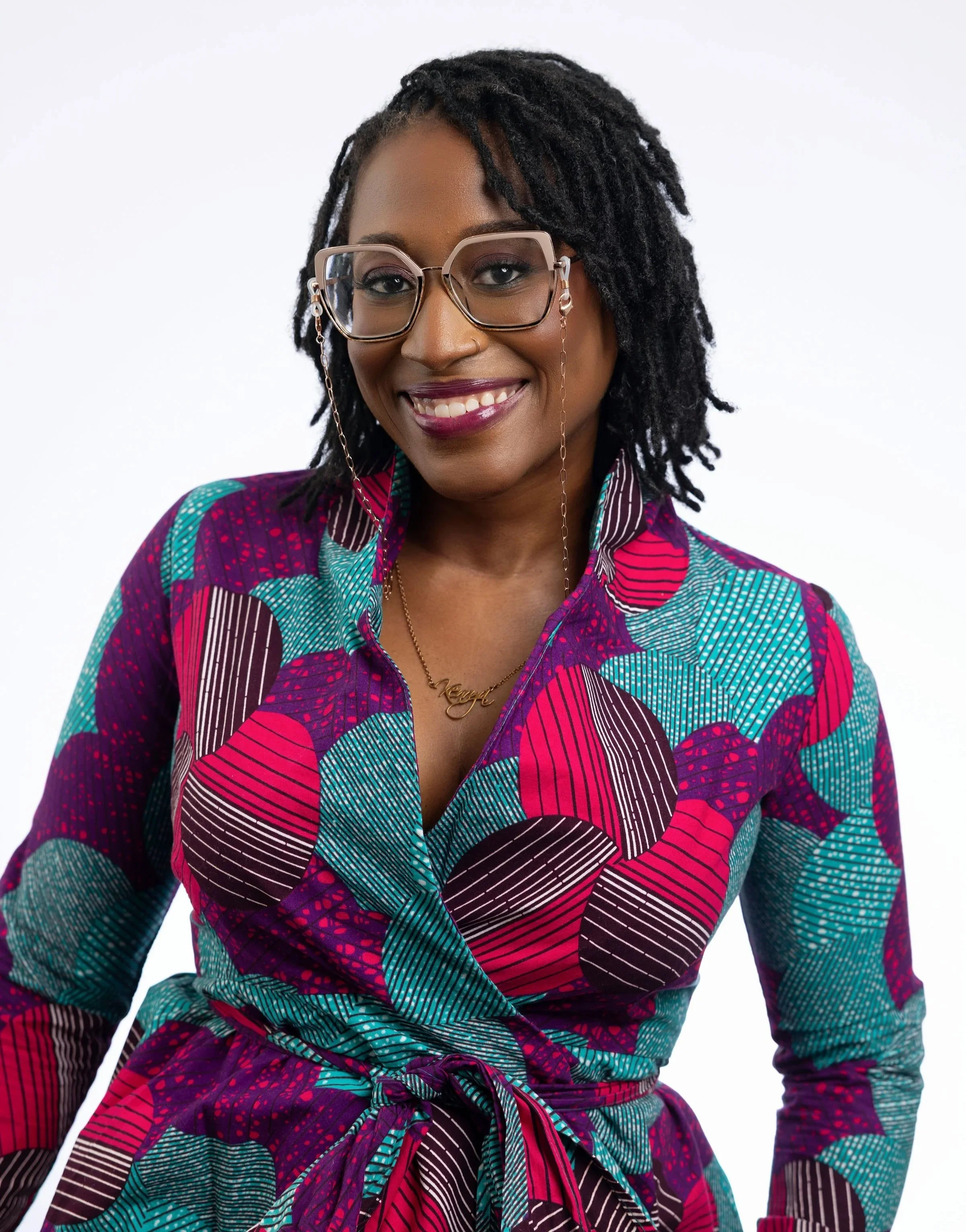 This is Kenrya Rankin, a dark-skinned Black woman with dark brown locs, wearing rose gold glasses on a chain, purple lipstick, and a big smile. She is also wearing a shirt with a hot pink, purple, and teal print. The background is white.