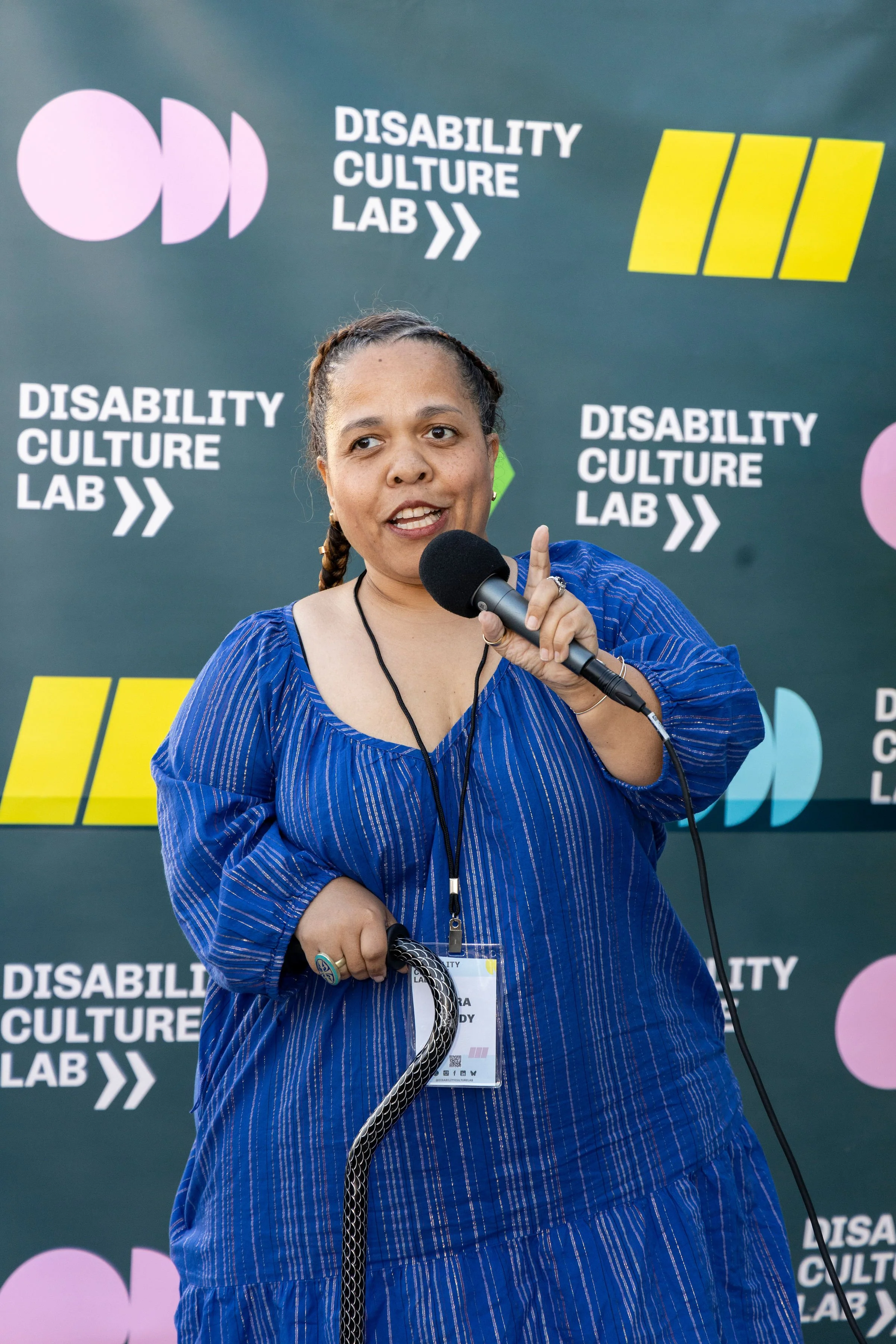 A Black woman of short stature wearing a striped Blue dress and holding a black cane speaks into a microphone while gesturing with her left hand and looking at the audience.
