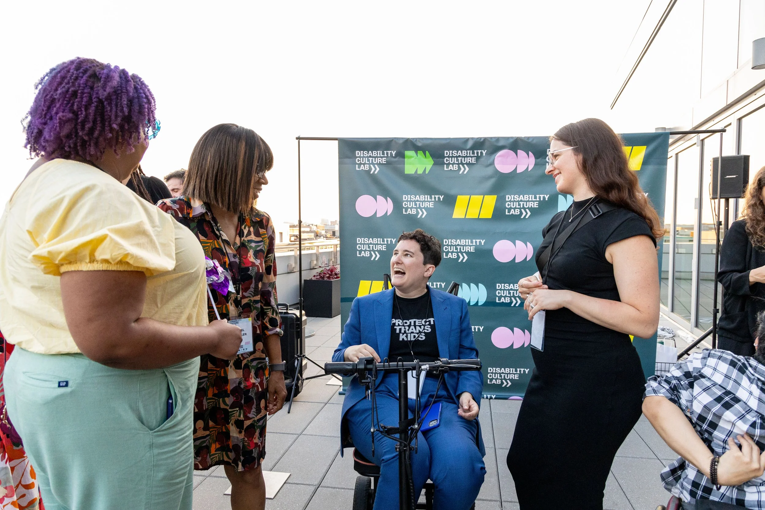 A diverse group of three women stand smiling and laughing while speaking to a person seated in a mobility device wearing a blue suit jacket and pants and a black t-shirt that reads "Protect Trans Kids."