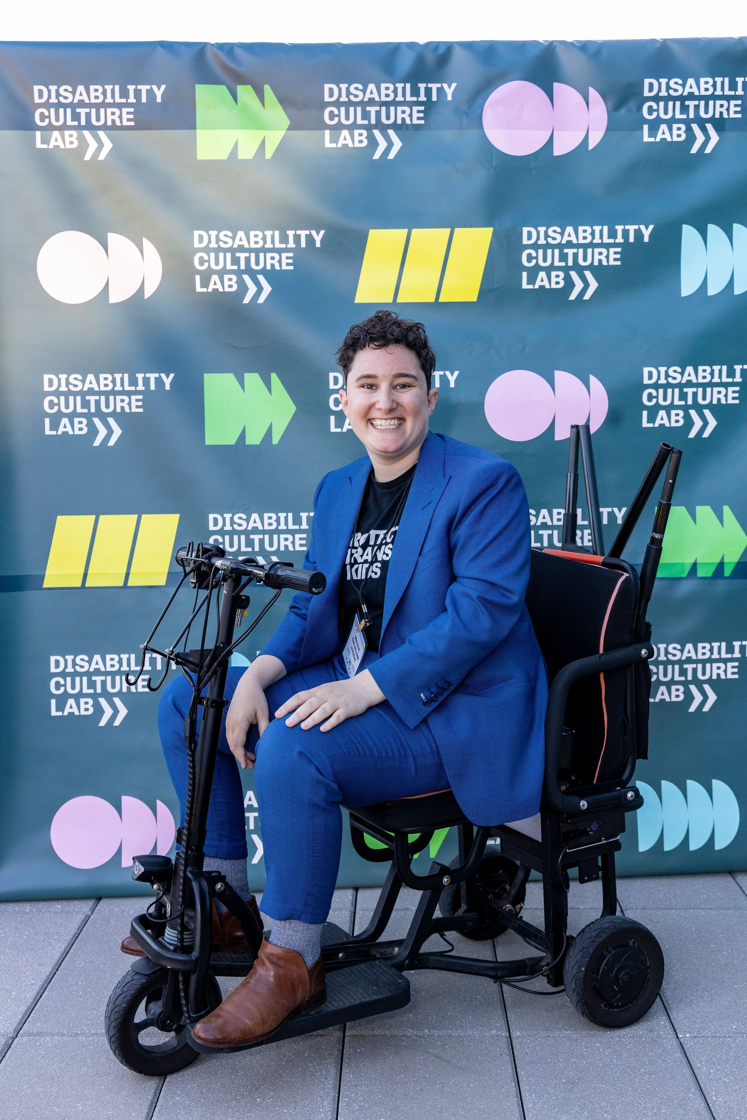 Disability Culture Lab Founder and Executive Director Meier Galblum Haigh sits on a mobility scooter in front of a “Disability Culture Lab” banner wearing a bright smile and a blue suit.