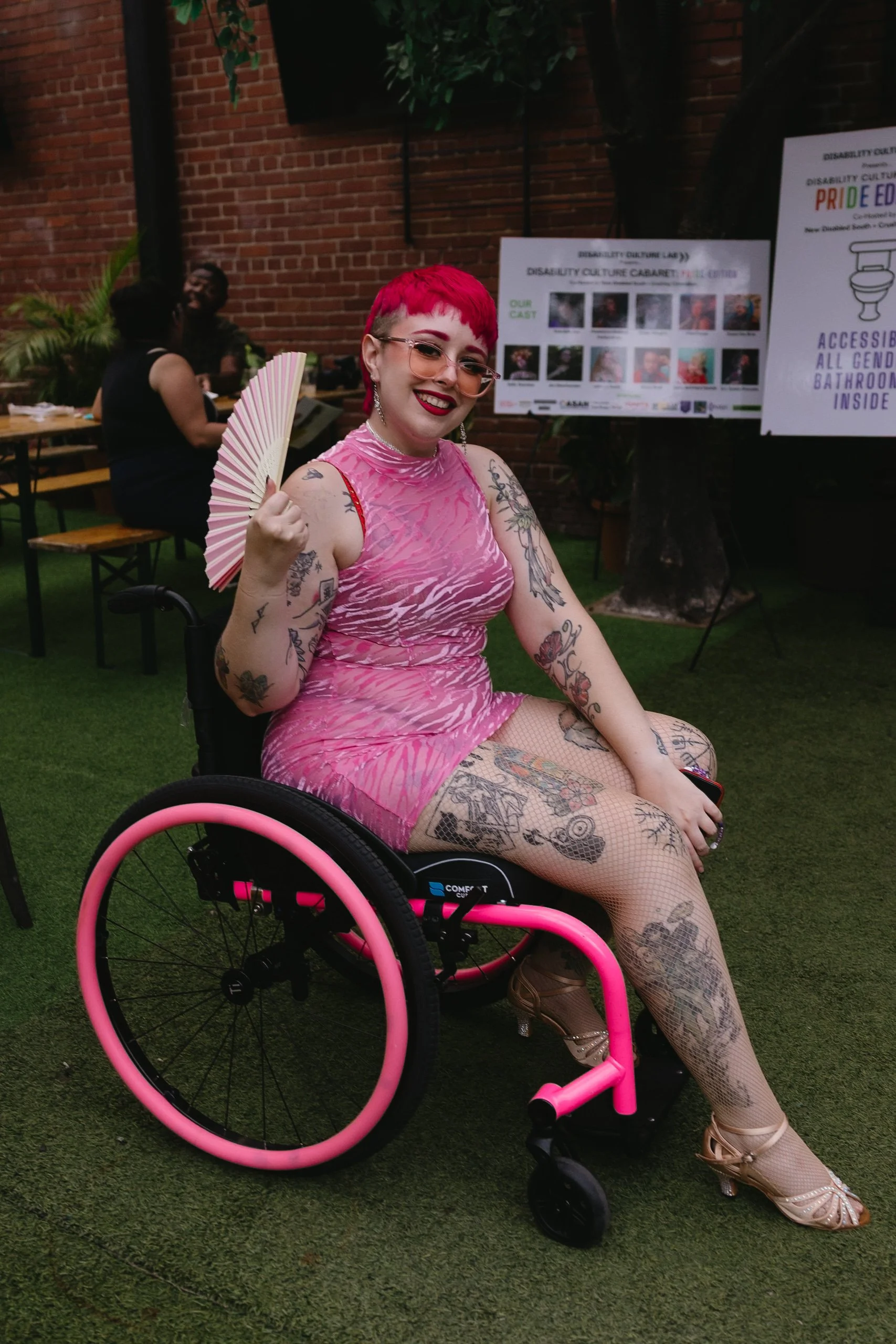 A smiling person with short pink hair, tattoos, and glasses wears a pink patterned dress. They sit in a wheelchair with bright pink rims and hold an open fan.