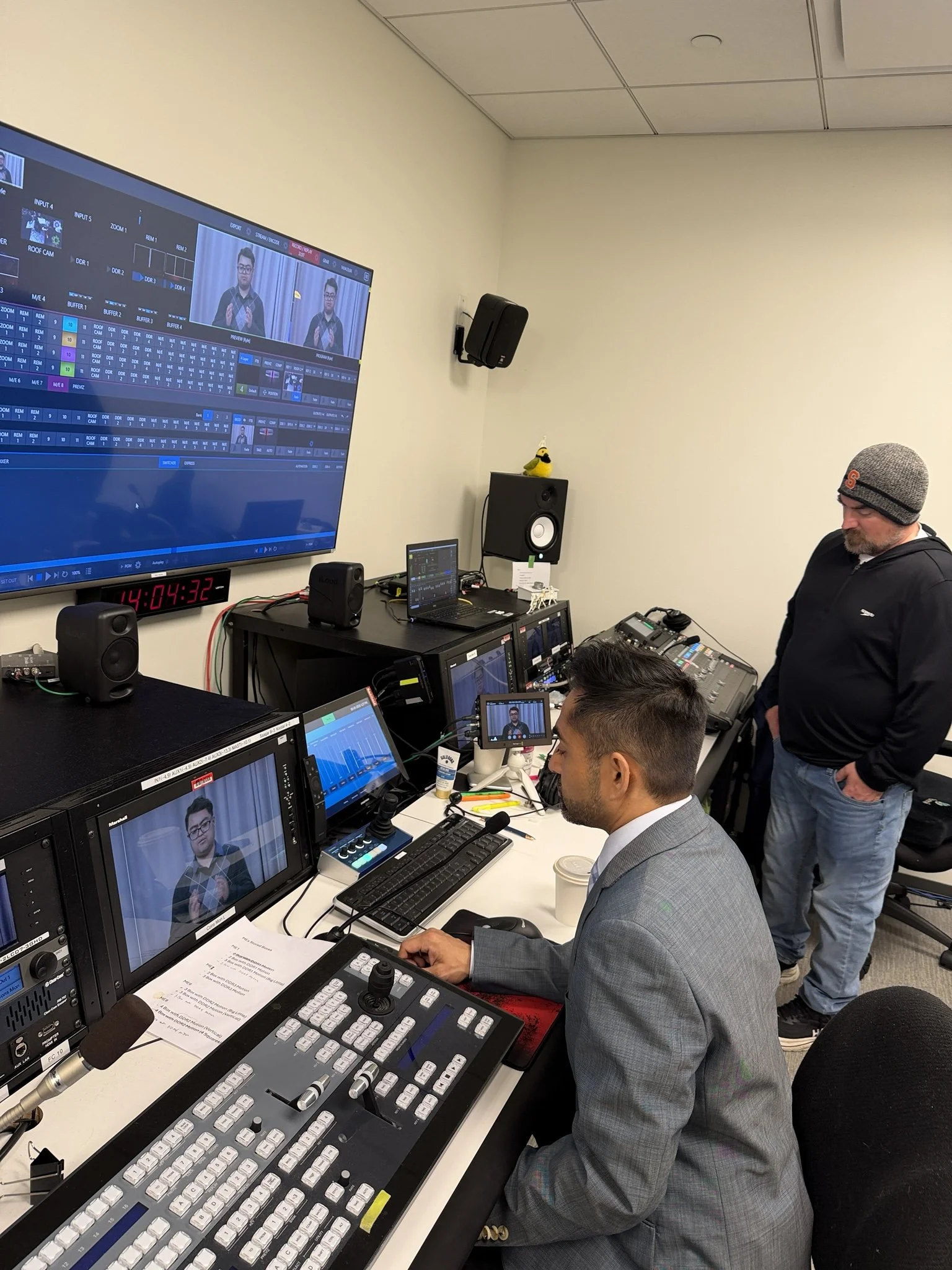 Two people monitor a live video production in a broadcast control room. One operates a video switcher while monitoring screens showing Disability Culture Lab Fellow Dennis Tran, and the other stands nearby observing.