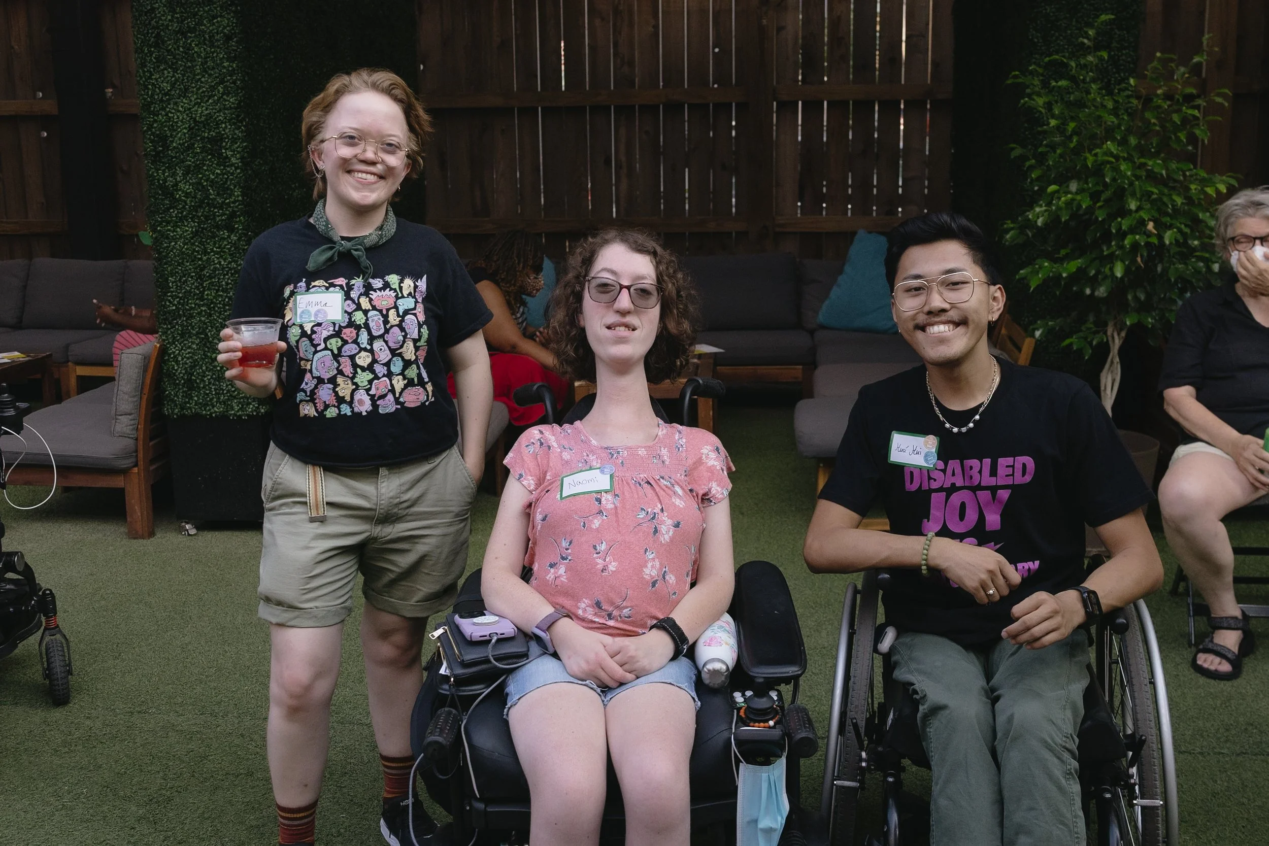 Three people smile together at an event. One stands holding a drink, while the other two sit in wheelchairs. They wear nametags and casual clothes, radiating a sense of joy and community.