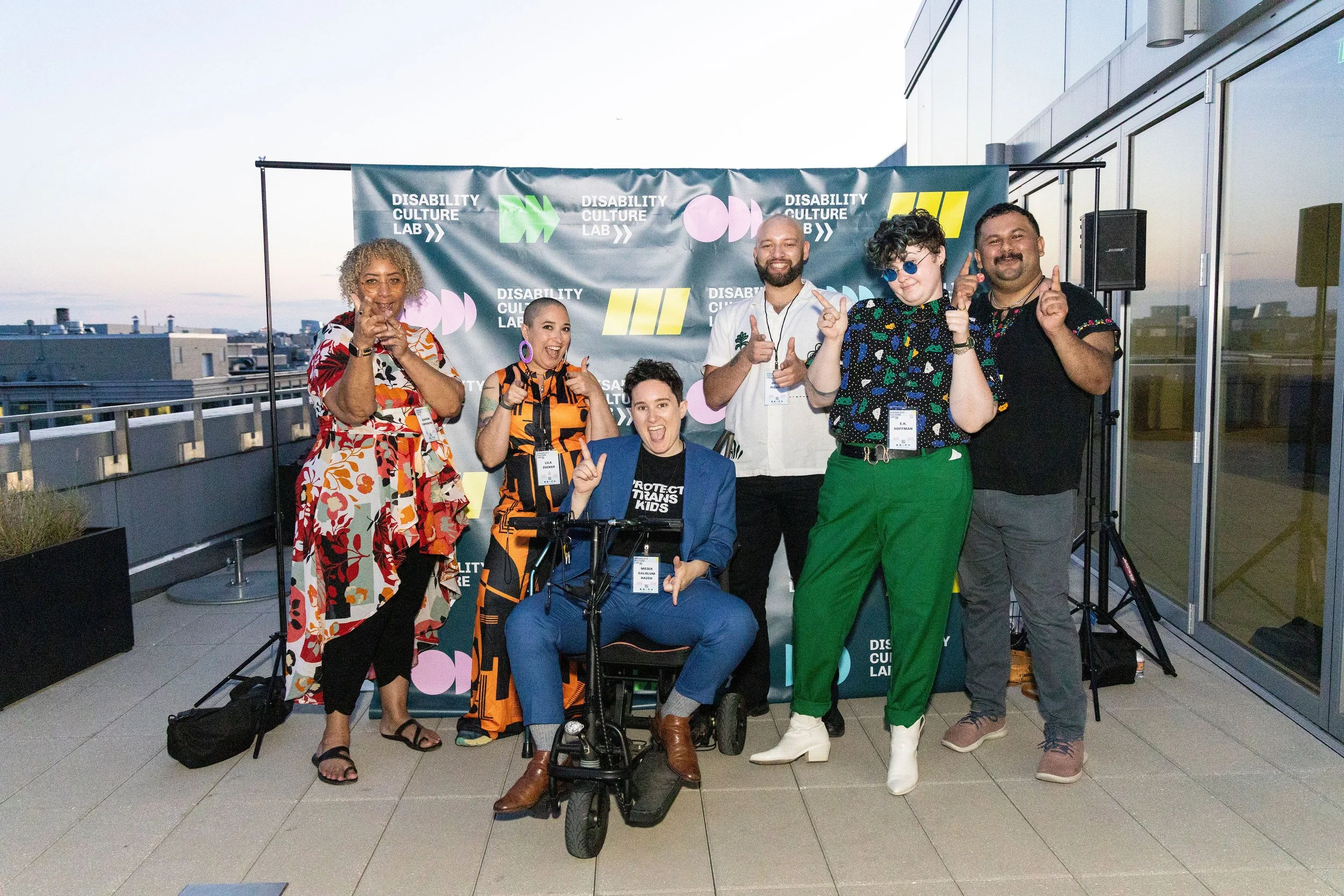 The Disability Culture Lab team poses together in front of a branded banner. One member sits on a scooter while others stand around them, all smiling and pointing playfully toward the camera.