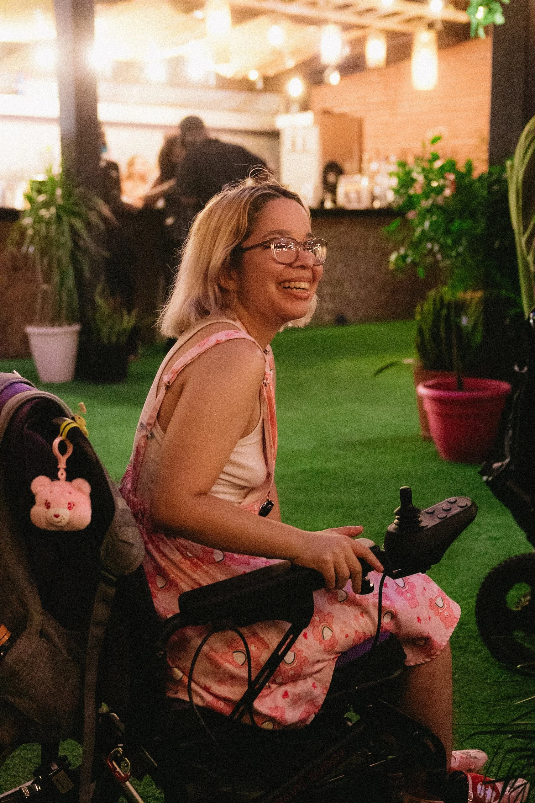 A woman with short blond hair and glasses smiles while seated in a power wheelchair at an evening event, wearing a pink dress in a warmly lit outdoor space.