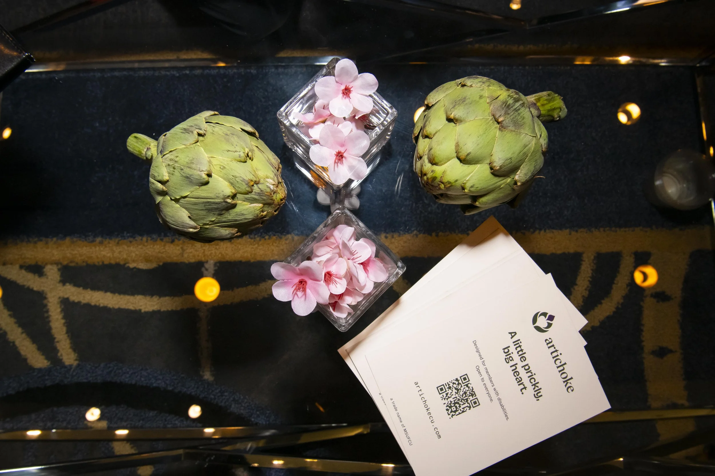 Two green artichokes sit on a glass table with small pink flowers in low rectangular vases and a card that reads, "Artichoke, A little prickly, big heart."