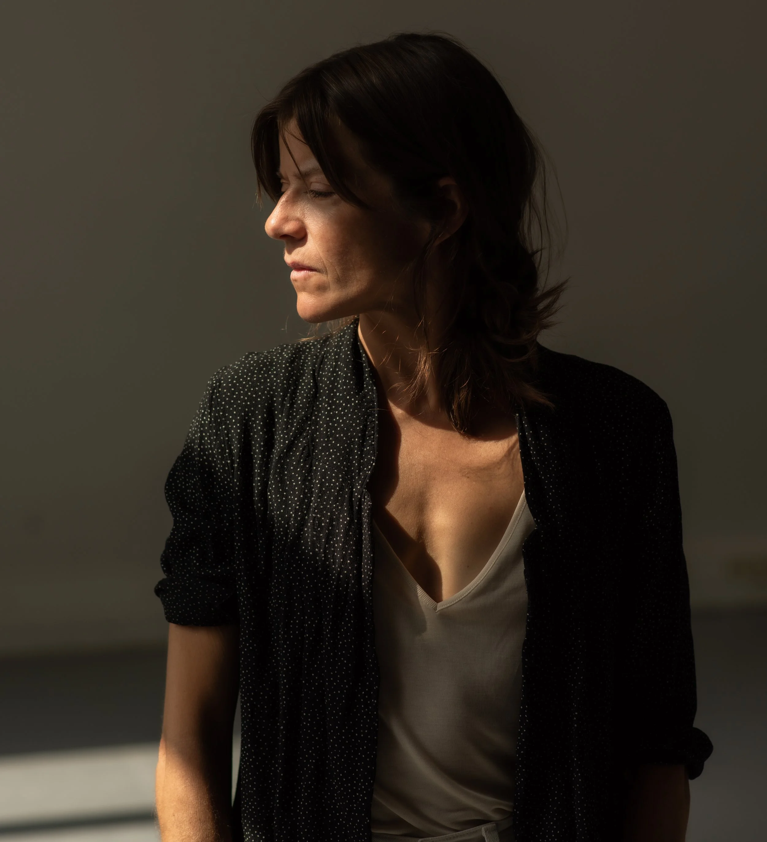 A woman with dark, shoulder-length hair looking down in a dimly lit room, wearing a black polka-dotted shirt and a beige top.