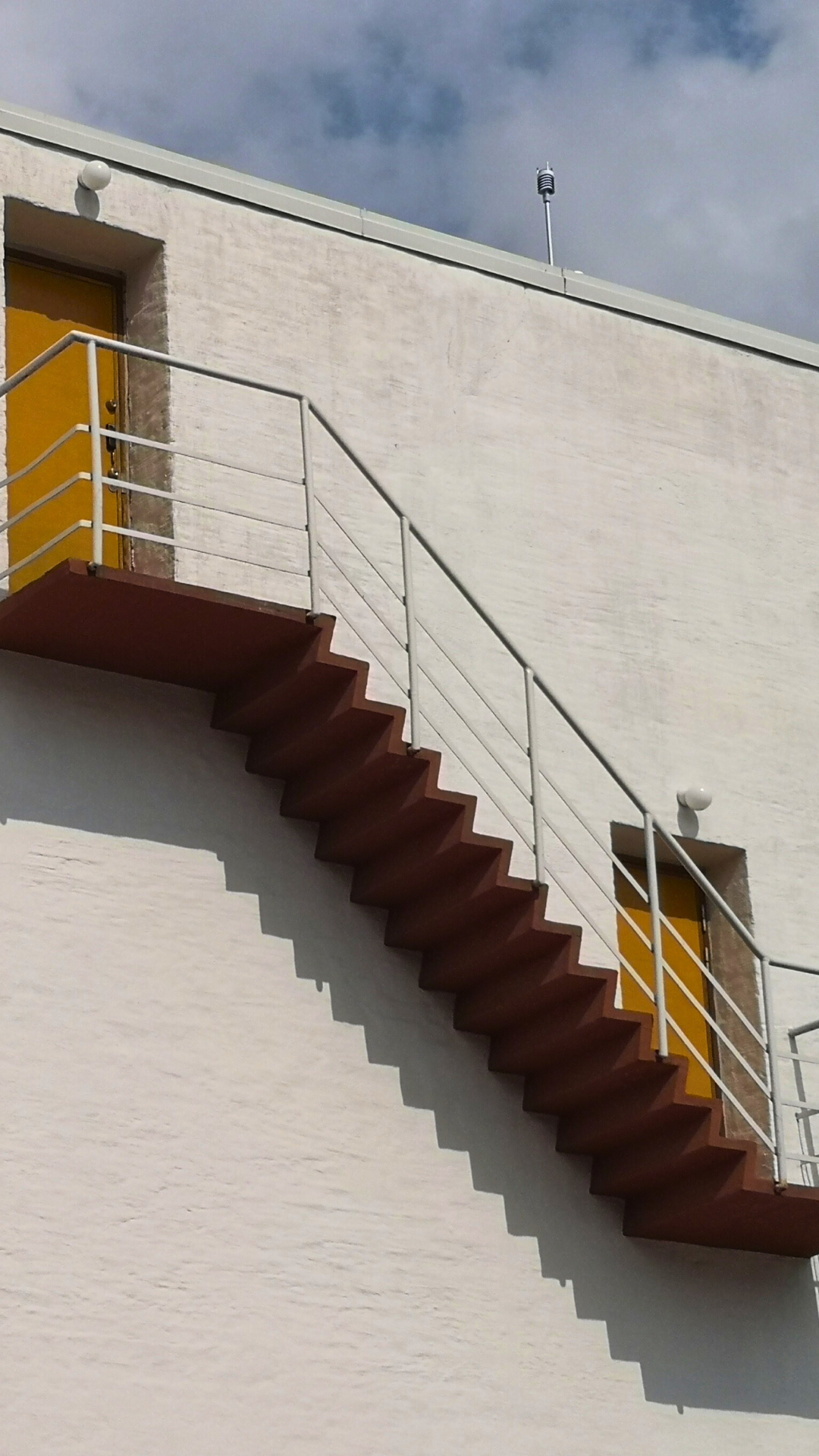 Exterior view of a white building with a staircase and yellow doors. The staircase has white metal railings and cast shadows on the wall. The sky is partly cloudy.