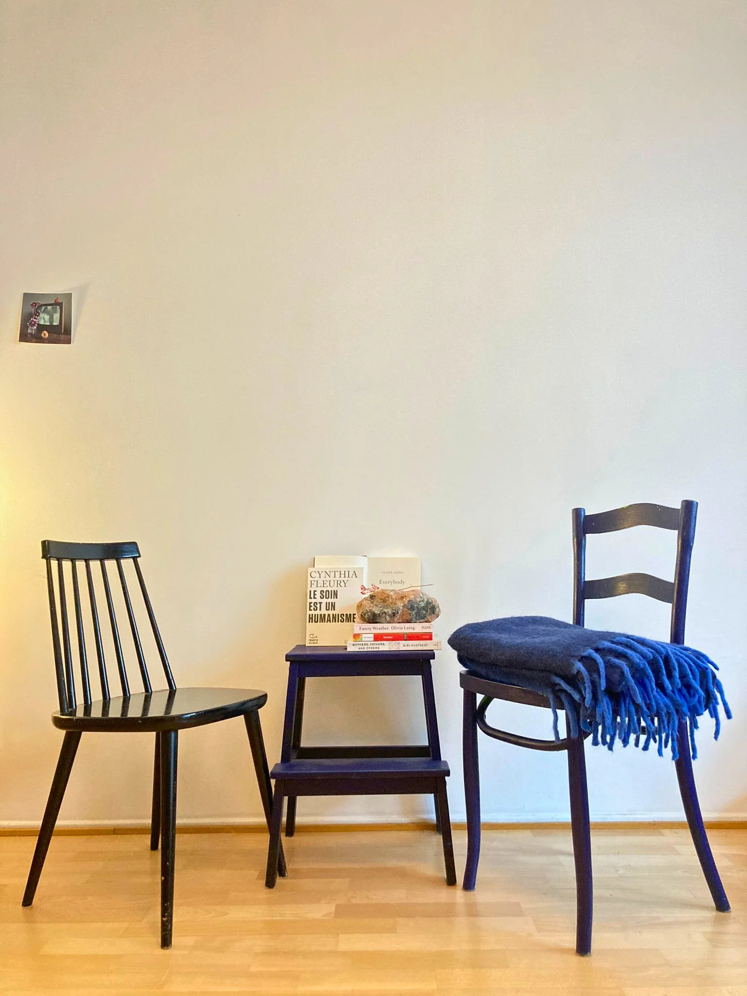 A plain white wall with a small framed picture on the left and a group of chairs arranged in a semi-circle. A small table with books, a rock, and a decorative item is in the center. One chair has a blue blanket draped over it.