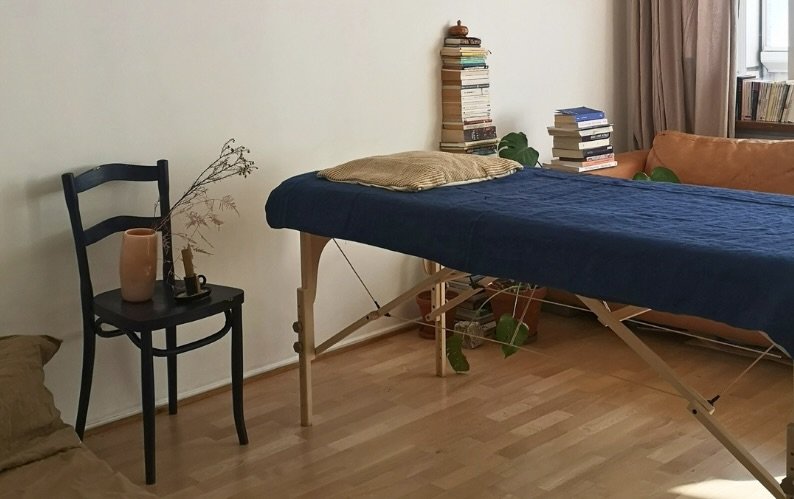 A room with a massage table covered in a dark blue sheet, surrounded by books, a chair with a vase and dried plant, a sofa, and window with curtains.