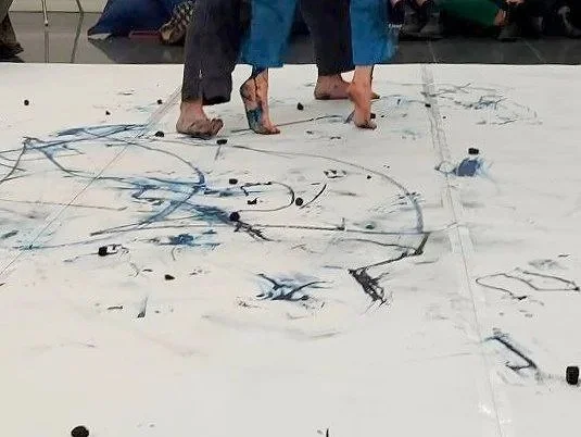 Two people with bare feet standing on a large white surface with black and blue paint splatters and lines, possibly an indoor sports court or art space.