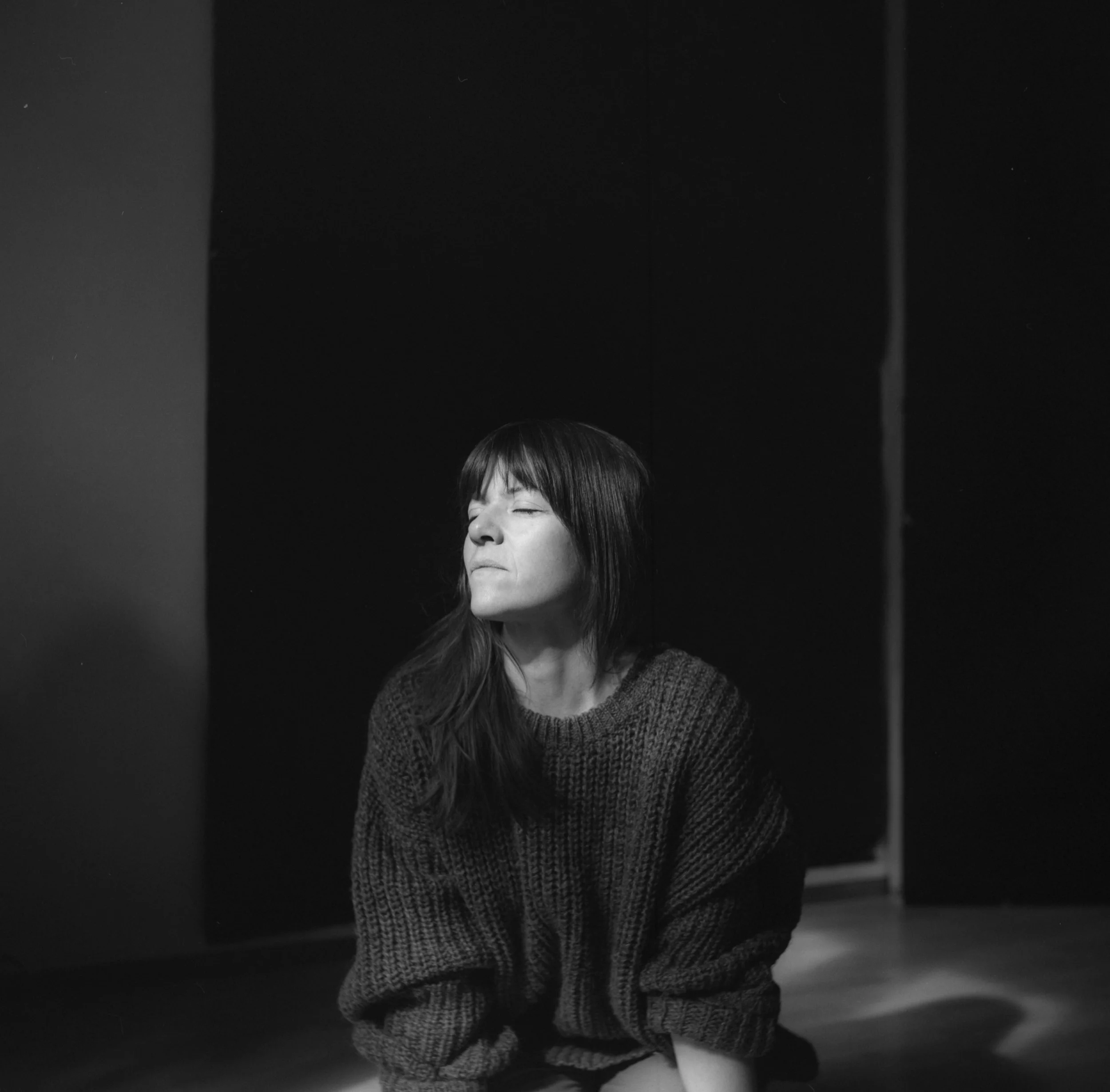 A woman with shoulder-length dark hair, closed eyes, and a content smile looking slightly to the side, wearing a textured sweater, indoors with dark wall in background.