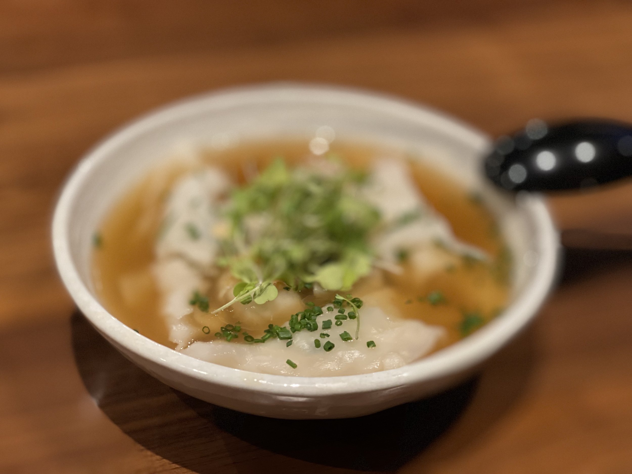 A bowl of dumplings in broth, garnished with chopped green onions and microgreens, on a wooden table, with a black spoon to the side.