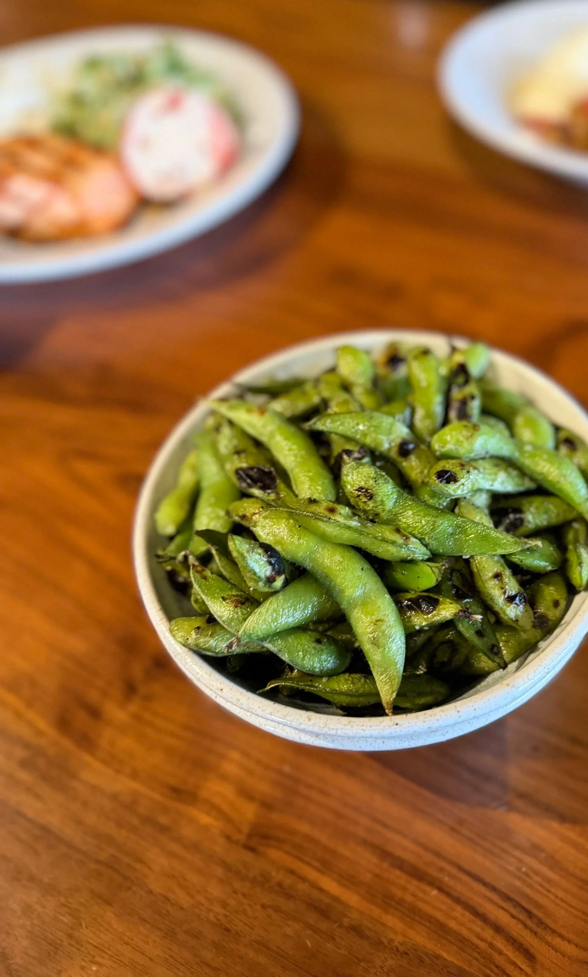 A bowl of grilled edamame beans on a wooden table with other dishes blurred in the background.