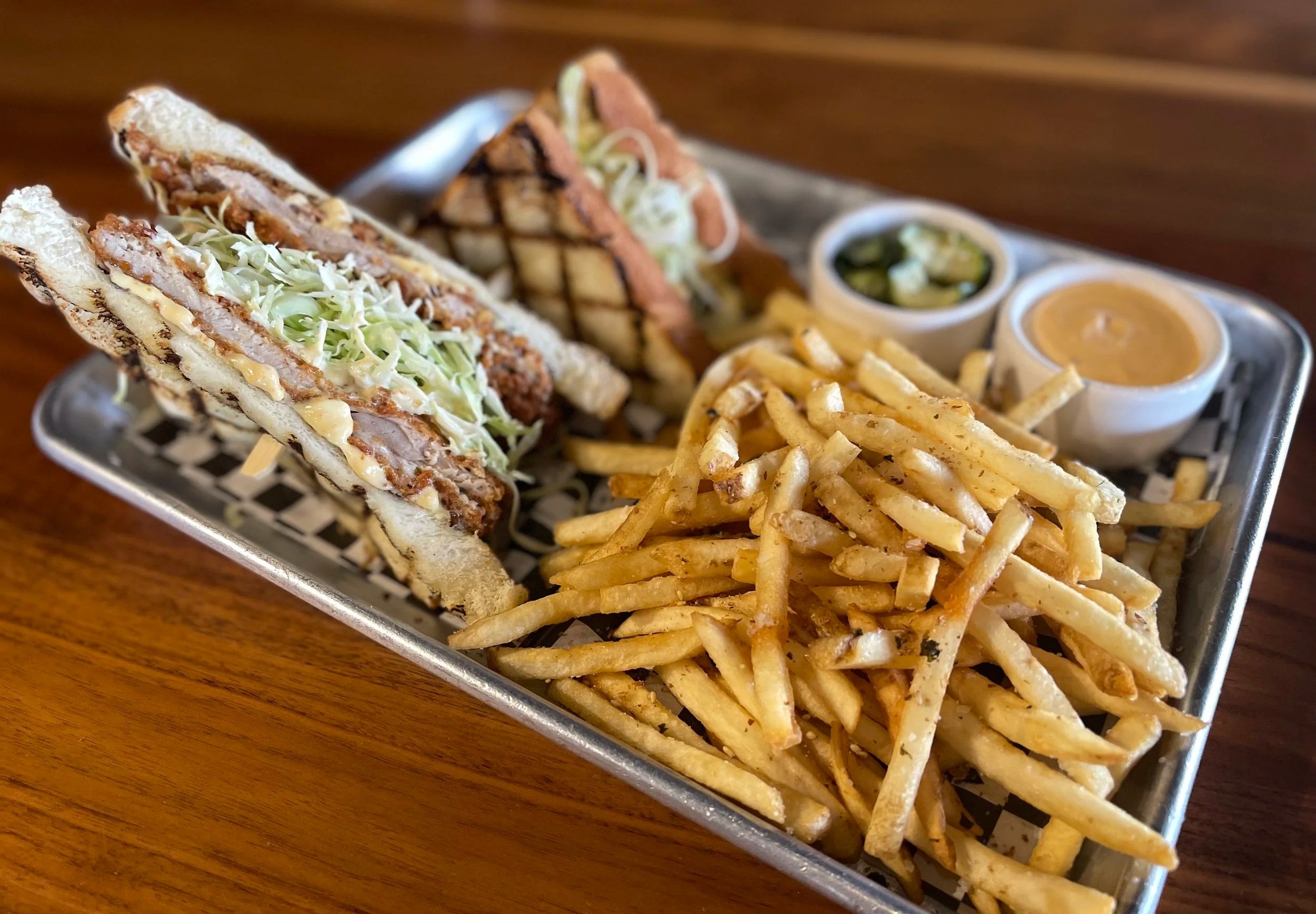 A metal tray with two clubs sandwiches filled with breaded meat, shredded lettuce, and sauce, served with a side of French fries, pickles, and two small cups of sauce on a wooden table.