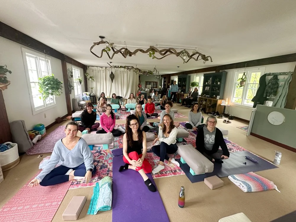 Group of women participating in a yoga or meditation class in a cozy, plant-decorated room with large windows and colorful rugs.
