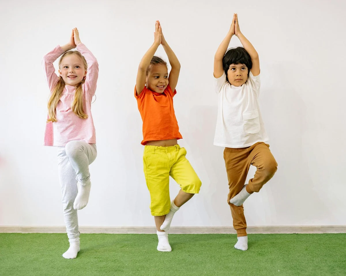 Three children practicing yoga, standing on one leg, with hands together above their heads, in a room with a white wall and green carpet.
