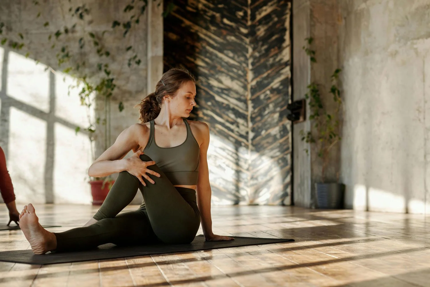 A woman practicing yoga indoors, sitting on a yoga mat, stretching her leg while holding her foot. The room has wooden floors and a textured wall with sunlight streaming through a window.