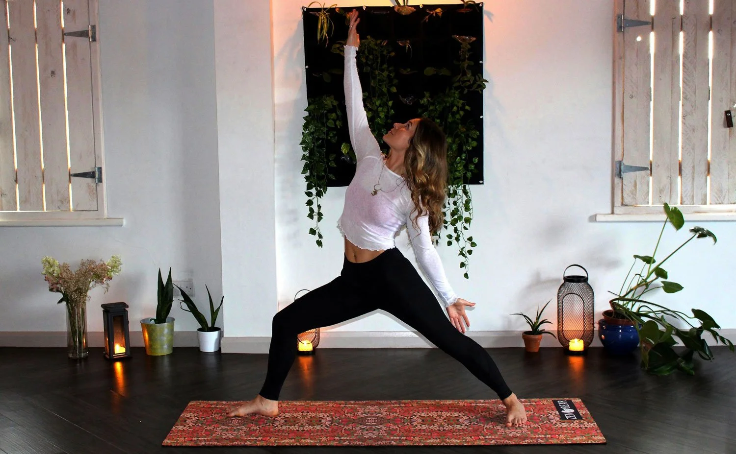 Woman practicing yoga in a room with plants, candles, and decorative lanterns, on a patterned yoga mat.