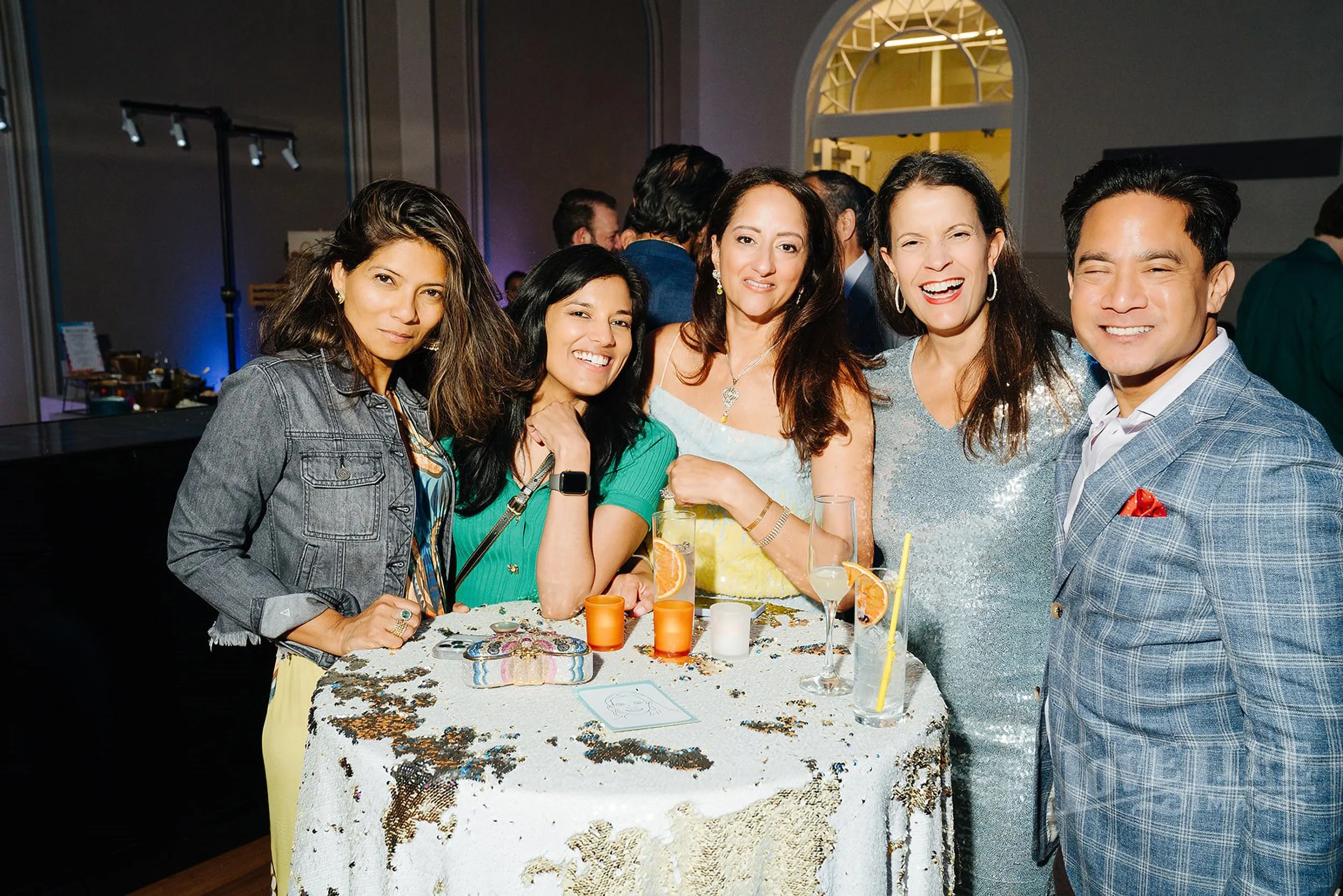 Elegantly-dressed party guests smile for the camera; cocktail table with sparkly linen is in front of them