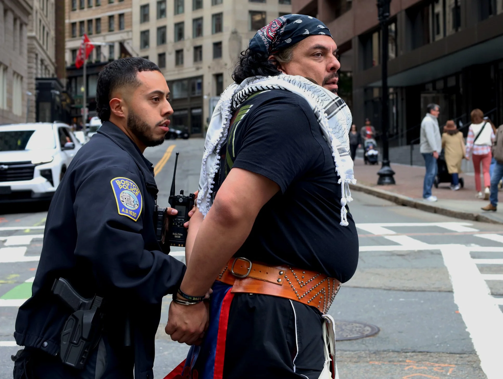 BOSTON, MA--10.11.25--Jean-Luc Pierite walks in handcuffs after being arrested by a Boston Police officer during Indigenous Peoples' Day March at the corner of Beacon Street and Tremont Street. 