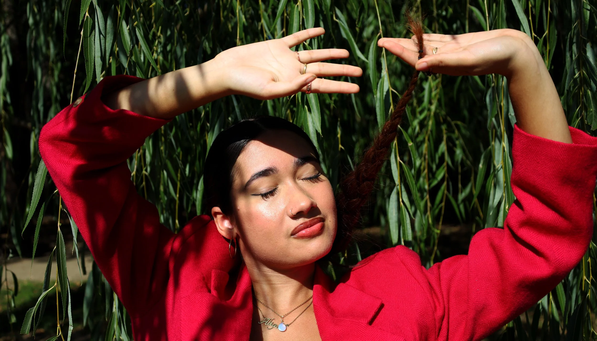 BOSTON, MA--10.4.25--Abigail Rosario lifts her hands over her head with her braid, and closes her eyes under a willow tree at Amory Park. She wears a bright red blazer and jewelry.