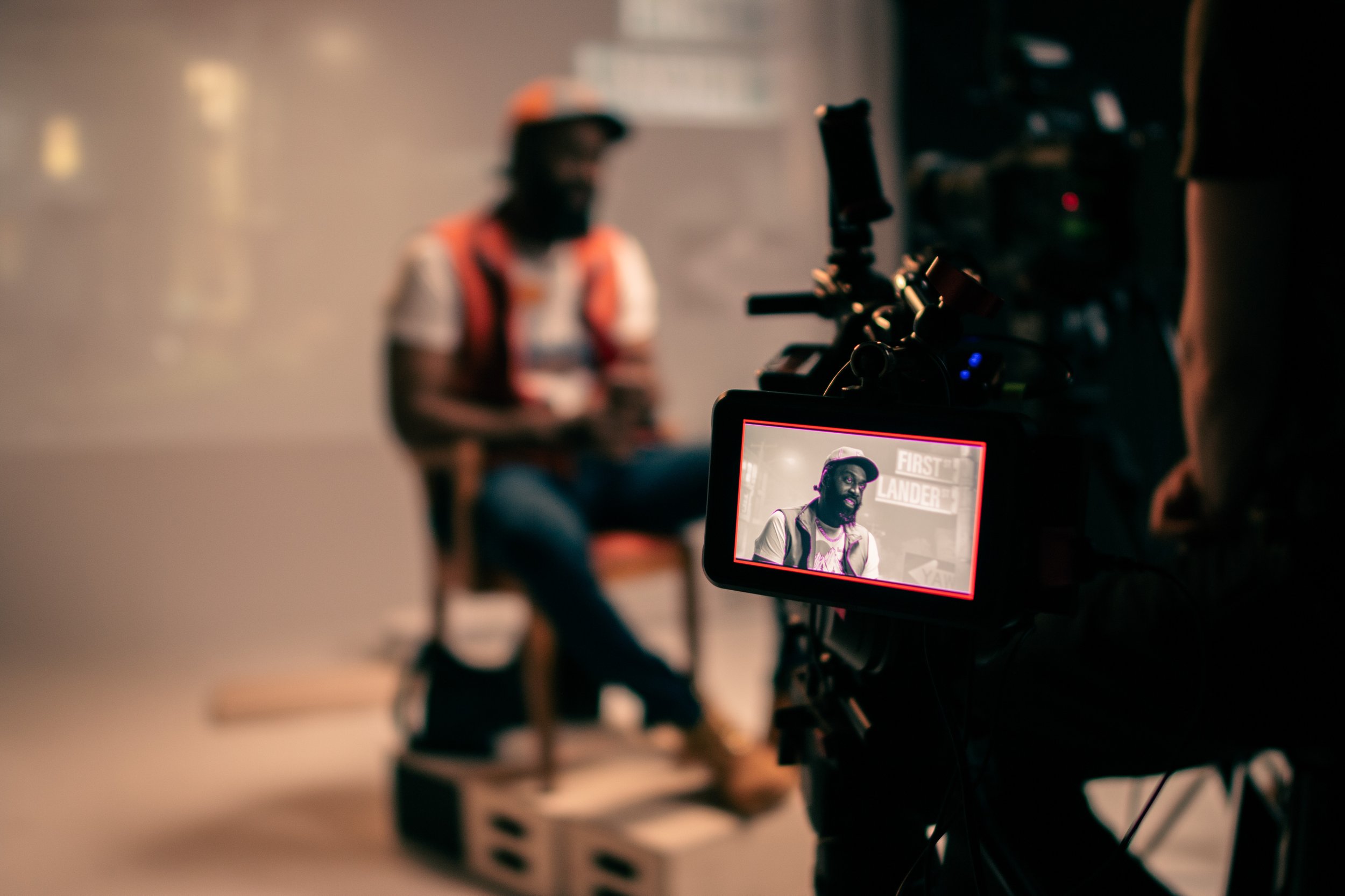 Behind-the-scenes view of a video interview shoot. A camera screen shows a person sitting and talking, with filming equipment visible.