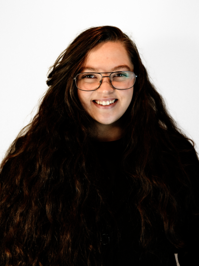 A woman with blonde hair wearing a black sweater, colorful plaid scarf, blue jeans, and brown boots holding a white heron sculpture in her hands, smiling, against a plain white background.