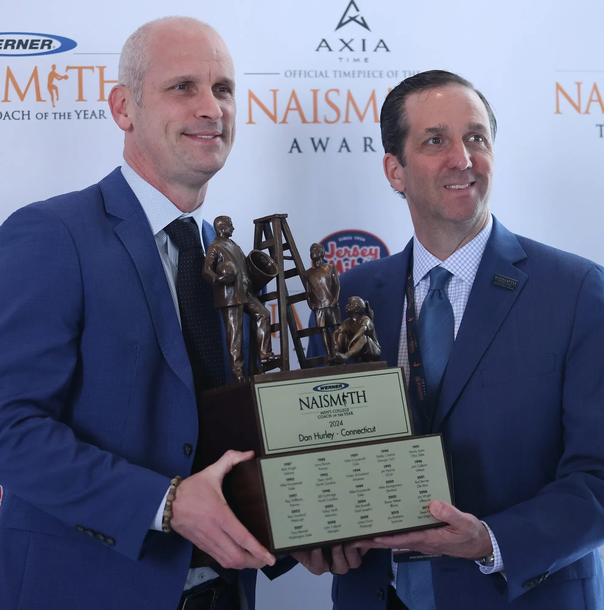 Two men in suits hold a Naismith trophy during a formal customs awards ceremony.