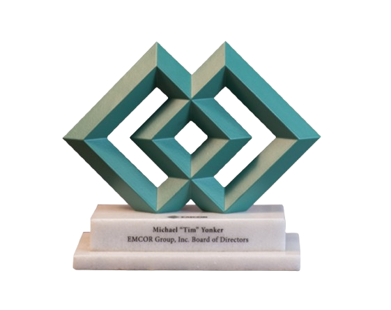 A geometric sculpture with a metallic finish resembling interconnected 3D shapes, mounted on a white marble base with a nameplate that reads 'Michael 'Tim' Yonker, EMCOR Group, Inc. Board of Directors'.