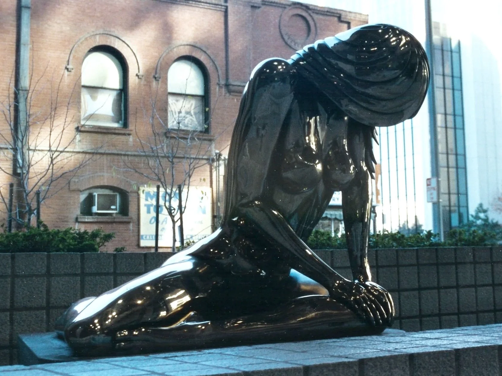 Black reflective sculpture of a woman kneeling with her head bowed, in an outdoor urban setting with brick buildings and leafless trees in the background.