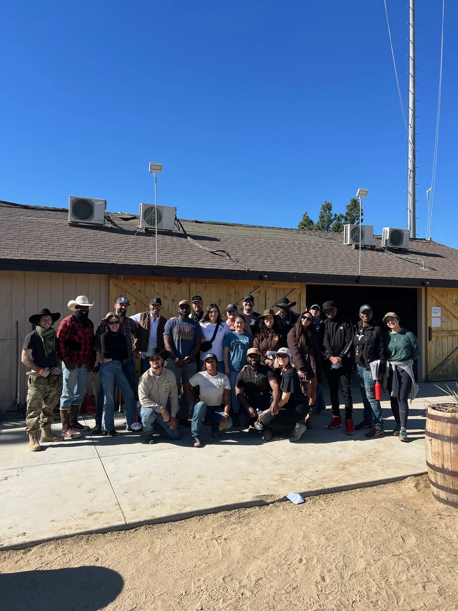 A group of people posing outdoors in front of a wooden building with a barn-style double door, under a bright blue sky.