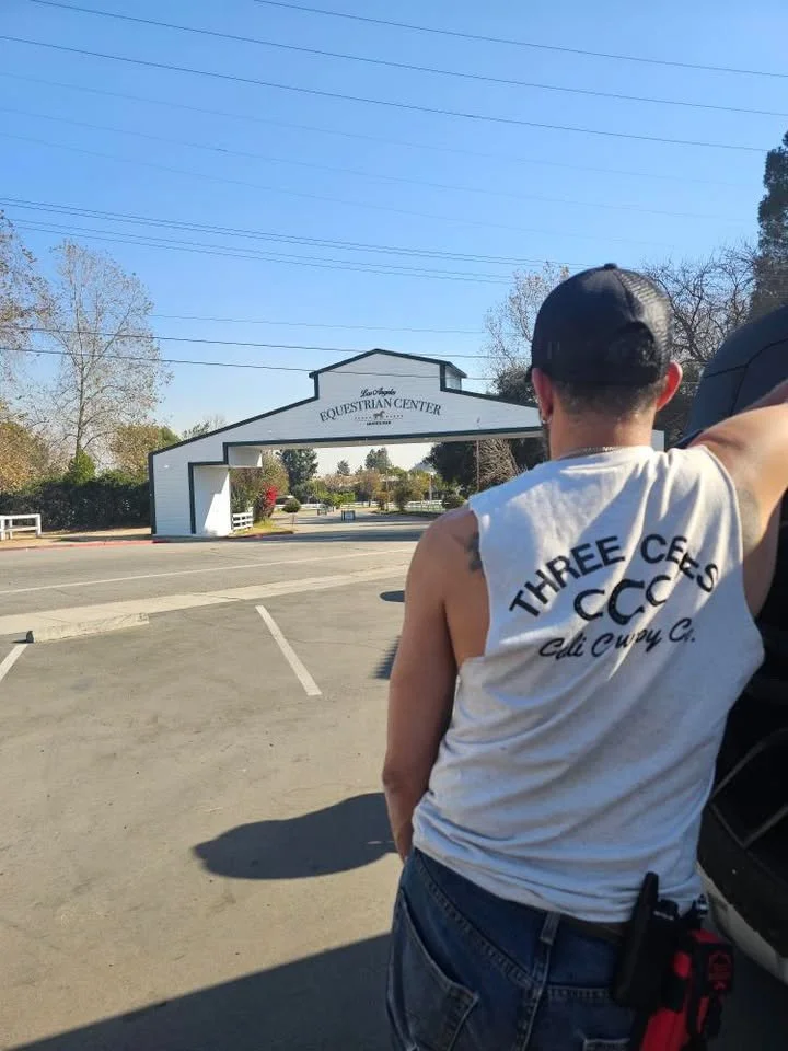 A man with a black cap and a sleeveless white shirt with the text 'Three Cee's Cali Cues C.' leaning against a vehicle in a parking lot near the Los Angeles Equestrian Center entrance, which is visible in the background under a clear blue sky.