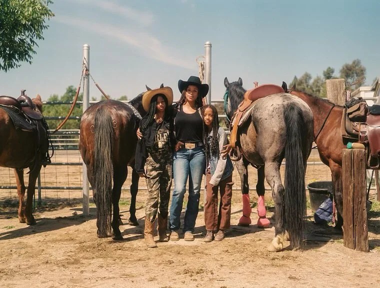 Four children and one adult standing with five horses at a ranch, some wearing hats, with a clear sky in the background.
