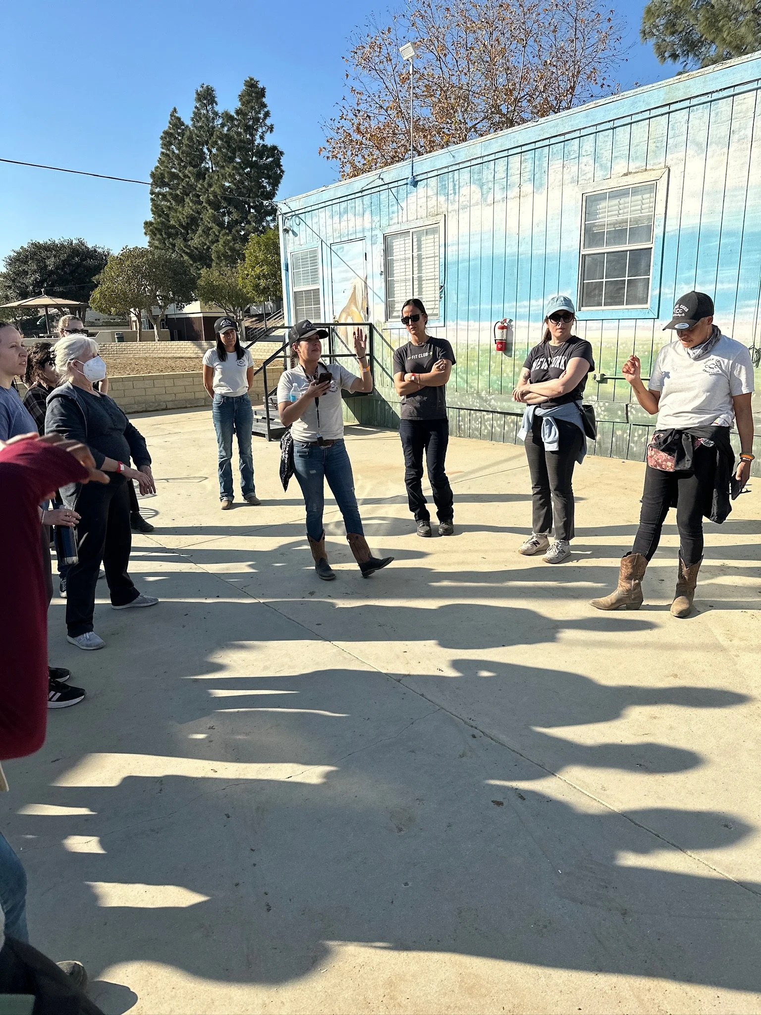 Group of people standing outside, some are listening to a woman who is speaking. Others are taking notes or listening, with a mural of a landscape painted on a building behind them. Some people are wearing hats and masks.