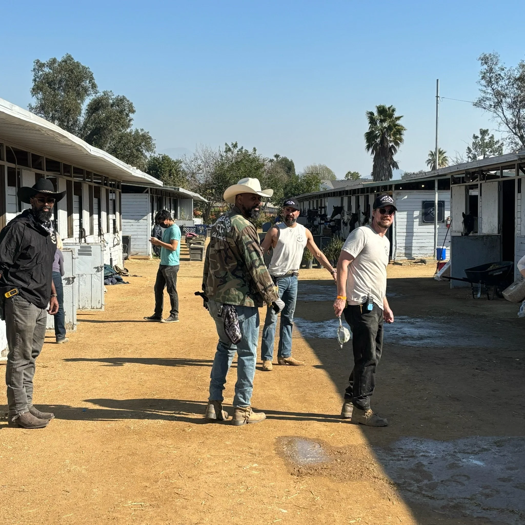 Group of people standing outside a horse stable, some wearing hats and sunglasses, with horses in the background on a sunny day.
