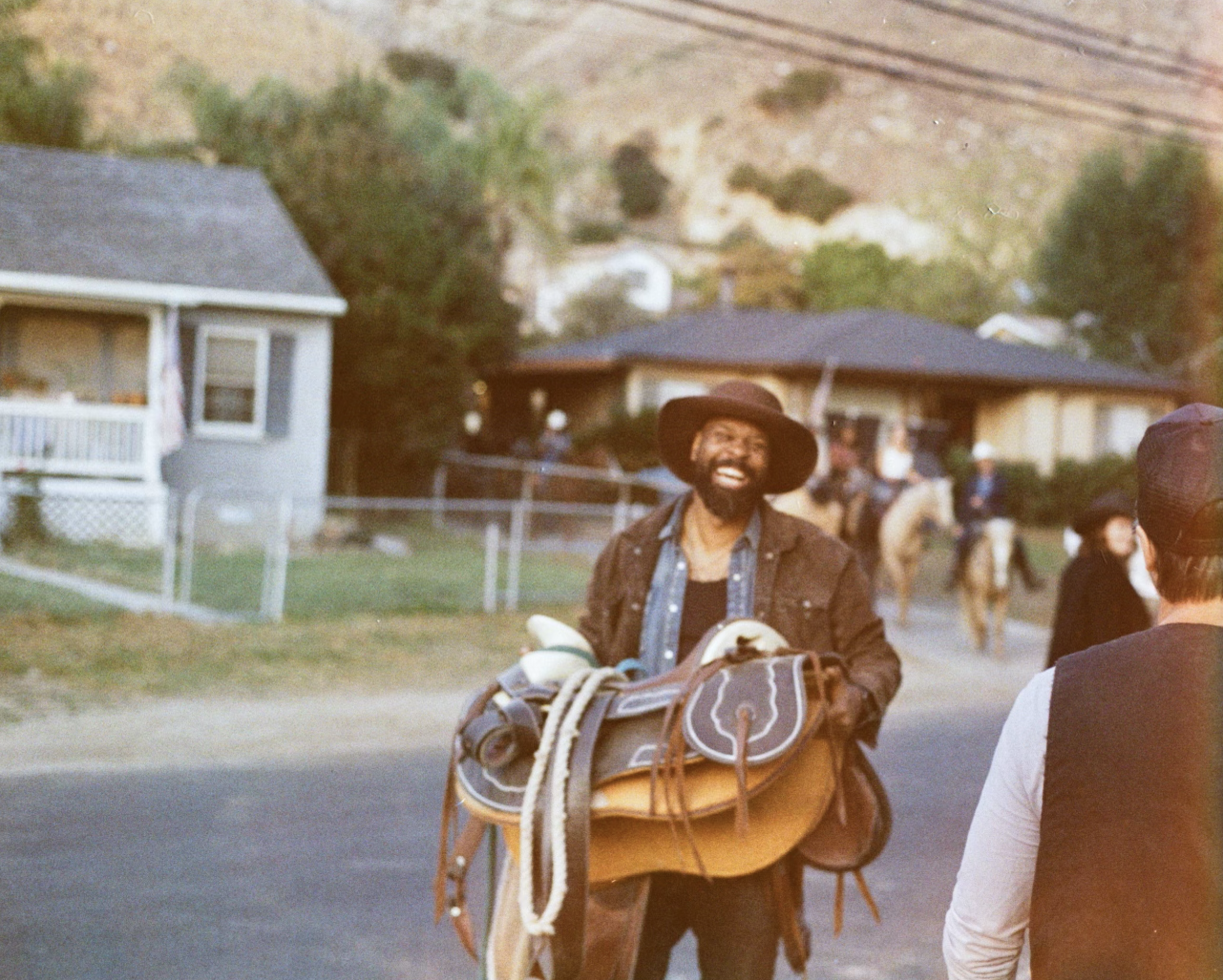 Man smiling and holding a saddle, carrying riding gear, wearing a large hat and a brown jacket, outdoors in a suburban neighborhood with other people in the background.