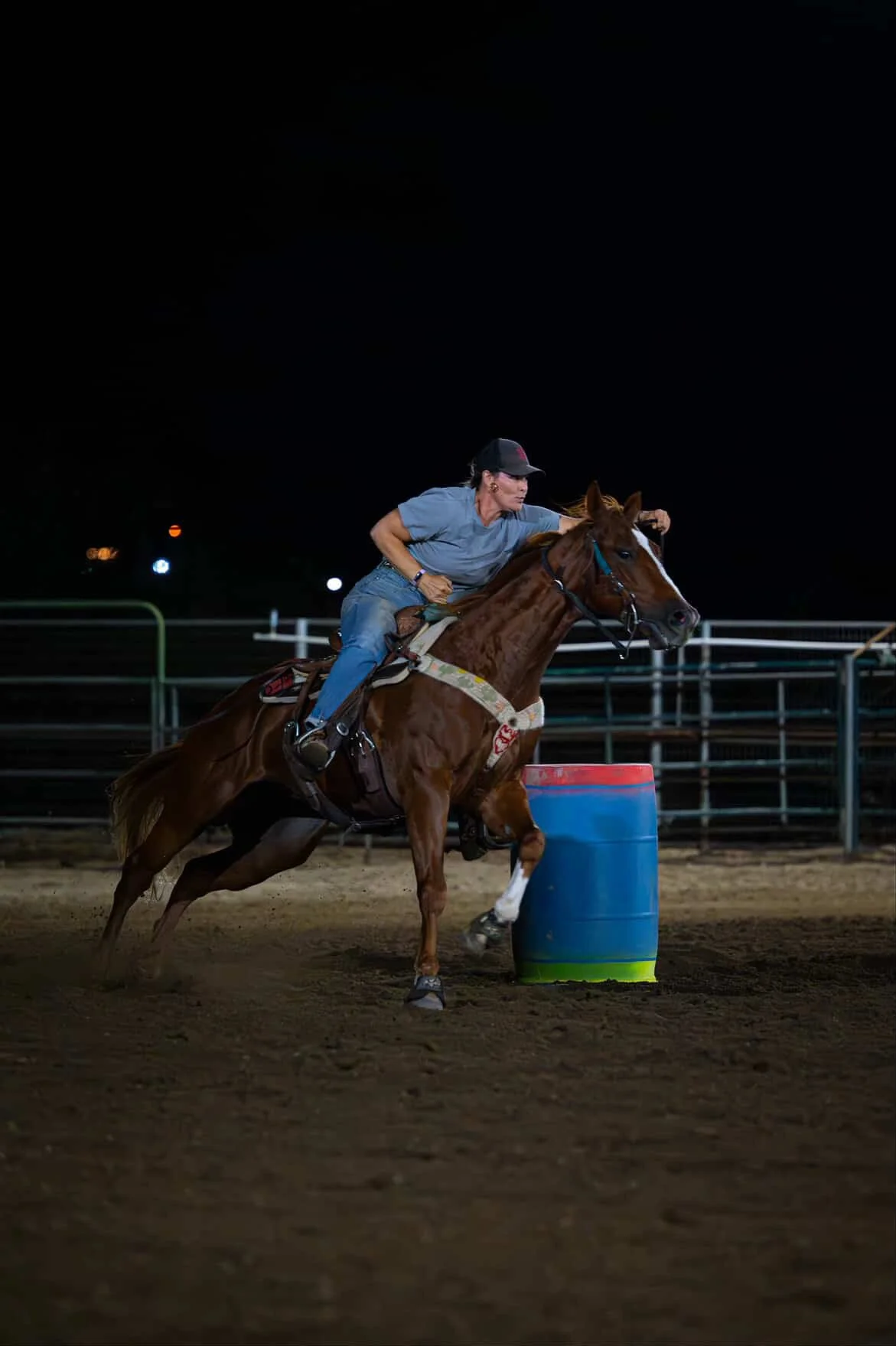 A person riding a horse at night, jumping over a blue and red barrel, in an outdoor arena with fencing.