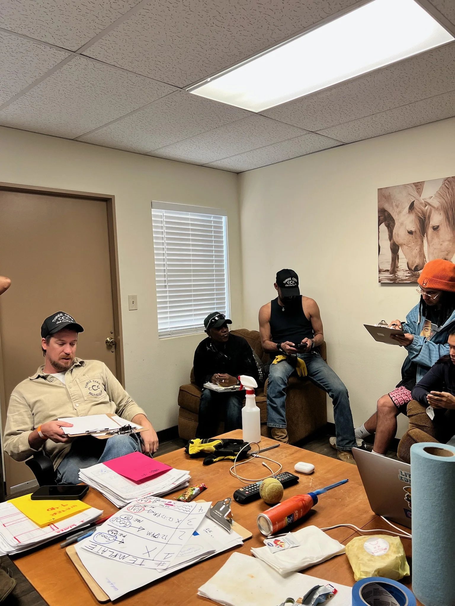 Group of people in a meeting room with cluttered table, some seated and others standing, with papers, office supplies, and a laptop, and a horse painting on the wall.