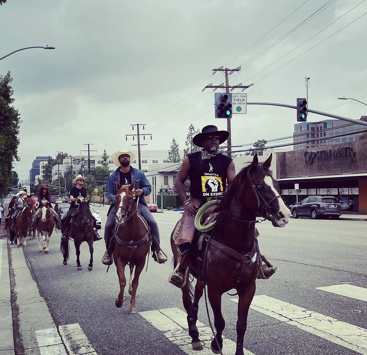 A group of people riding horses on a city street during a protest, with some wearing black shirts that say 'SAG-AFTRA ON STRIKE.'