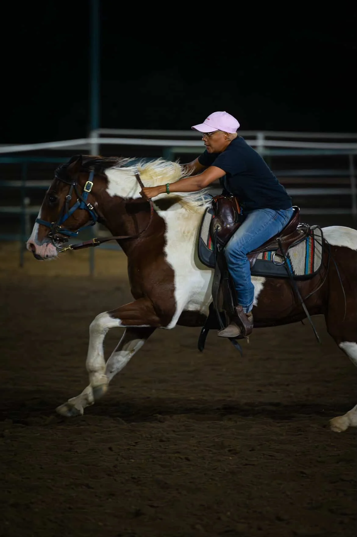 A woman riding a brown and white pinto horse at night on a dirt surface, with horse tack and a saddle.