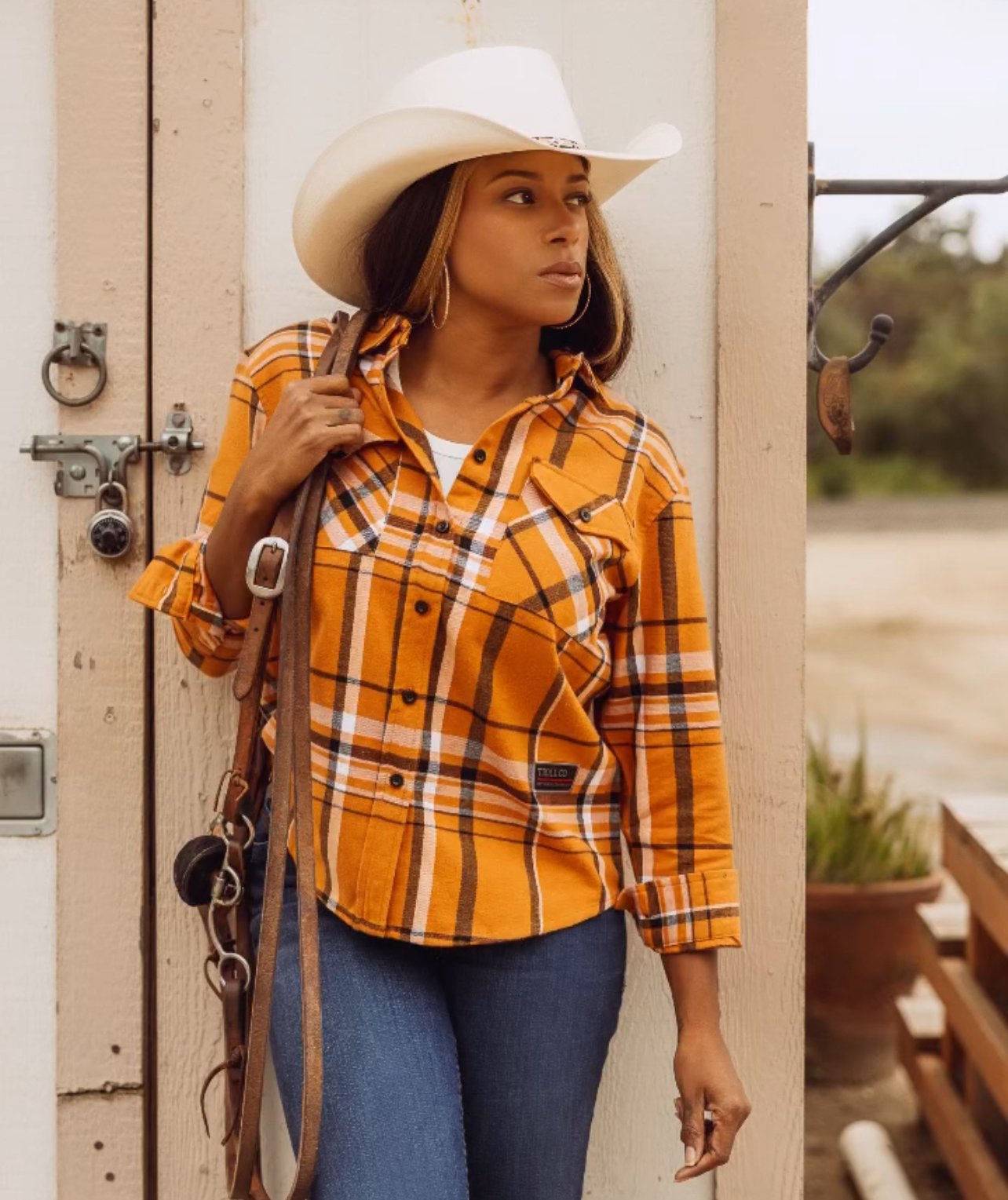 A woman wearing a white cowboy hat, orange plaid shirt, and blue jeans leaning against a wooden wall.
